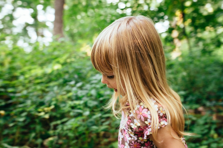 Young girl with blond hair standing outside with the green leaves all rounder.