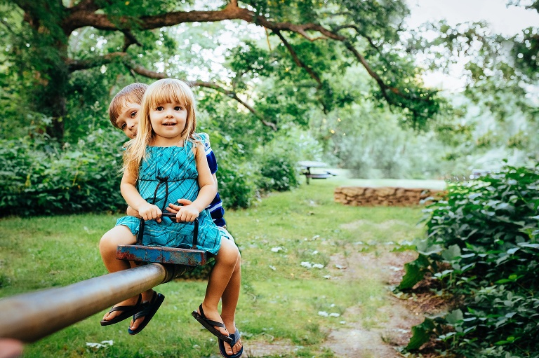 Young brother and sister sit on a teeter-toter together outside at a park.