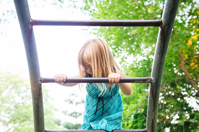 Young girl climbing up a ladder outside on a playground. Green trees behind her.