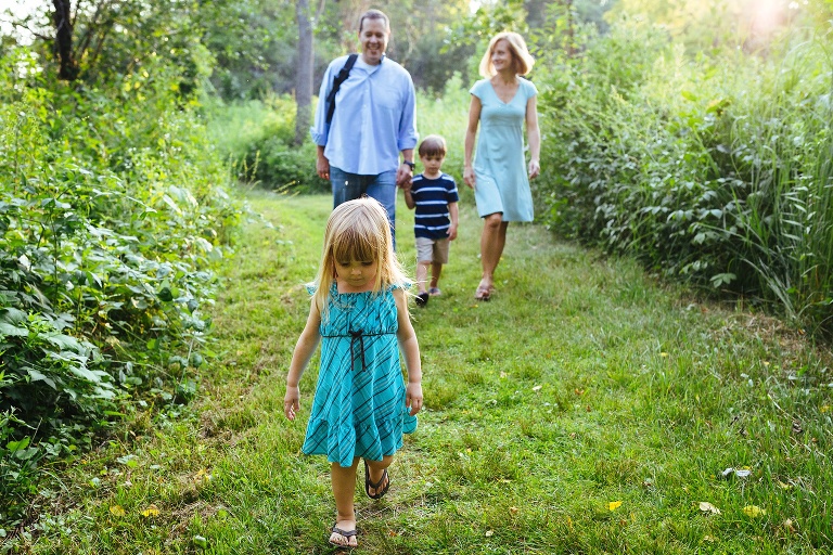 Family of four walking down a grassy path, young girl leading her parents and brother.