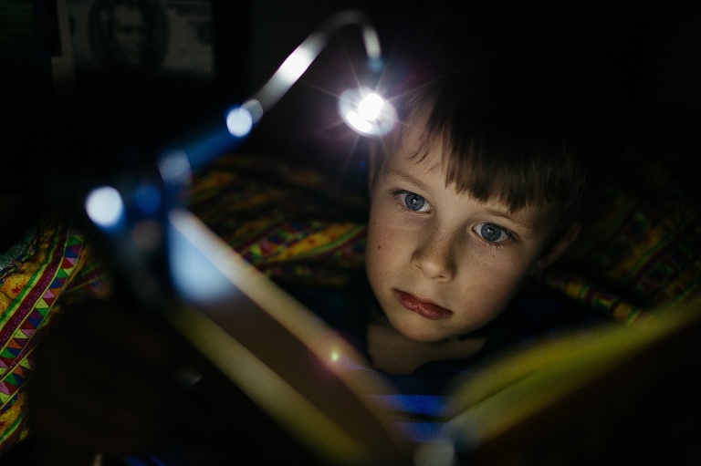 Boy sits in the dark reading a book with book light.