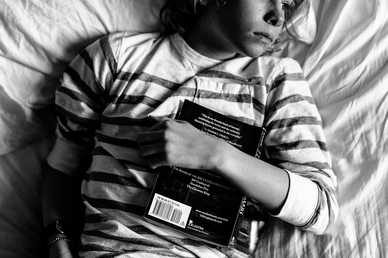 Black and white. Boy laying in bed with his arm wrapped around a book sitting on his chest looking out the window waiting for spring to come.