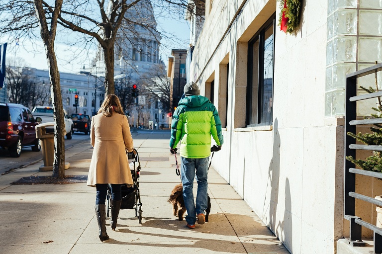 Mother pushes stroller as father walks two dogs down sidewalk downtown at christmas time.