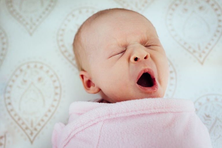 Newborn baby girl yawns as she's in swaddle blanket and lays in crib.