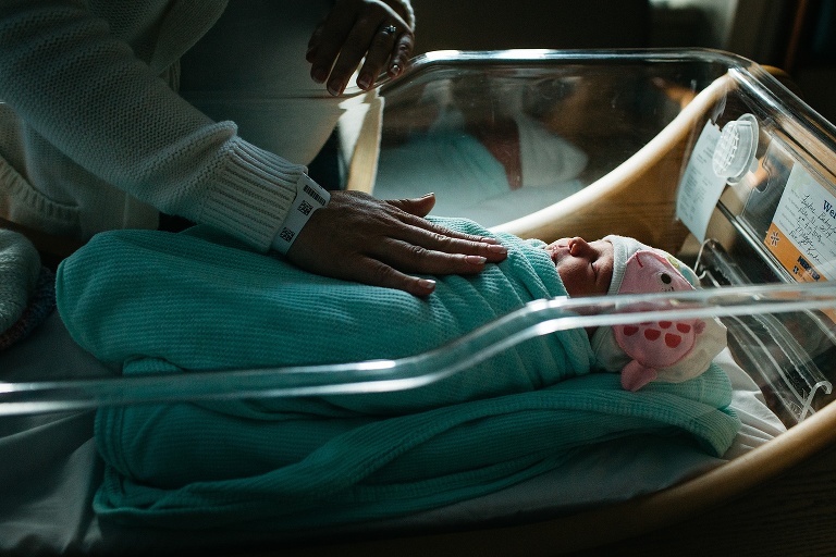 Sleeping newborn baby lays in bassinet in the hospital, mother watches and rests her hand on the baby.