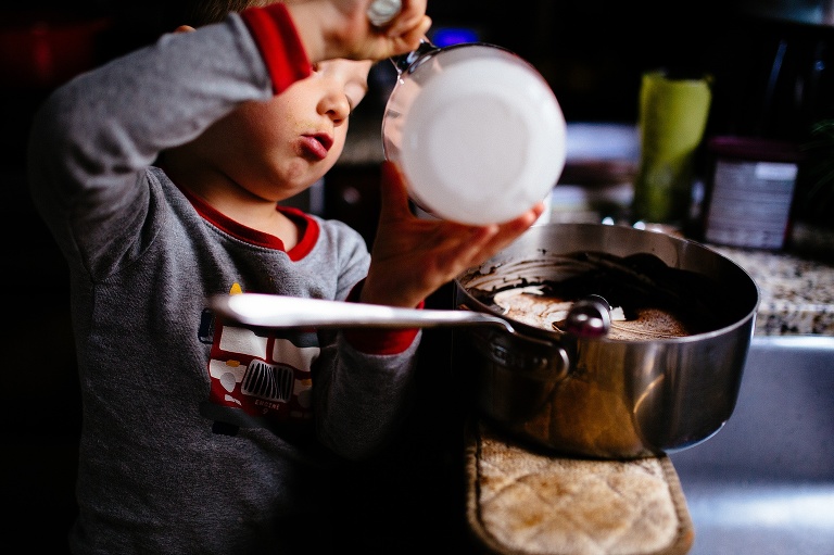 Toddler boy holds measuring cup of heavy cream and tips it into a silver saucer. 