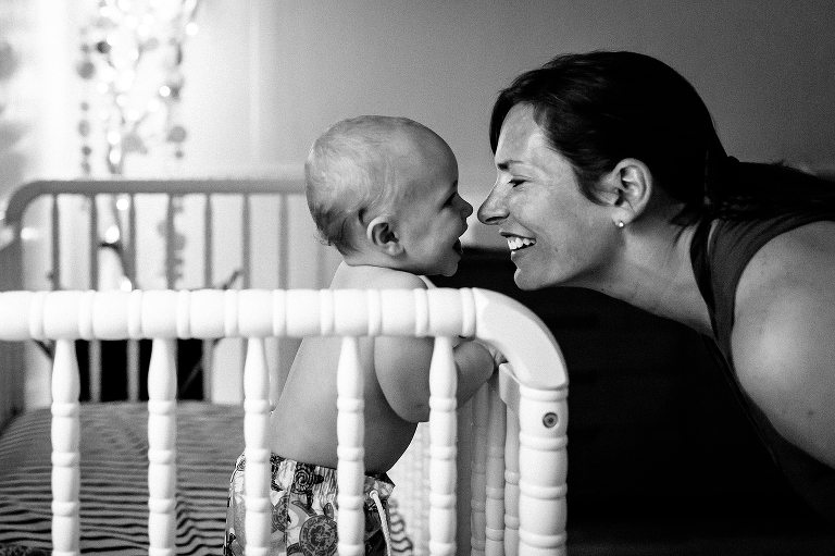 Black and white. Mom smiles nose to nose with toddler son in his crib.