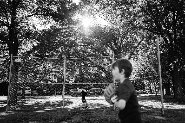 Black and white. Two boys play baseball together at local park. Many big tree's behind them with the sun shining through.