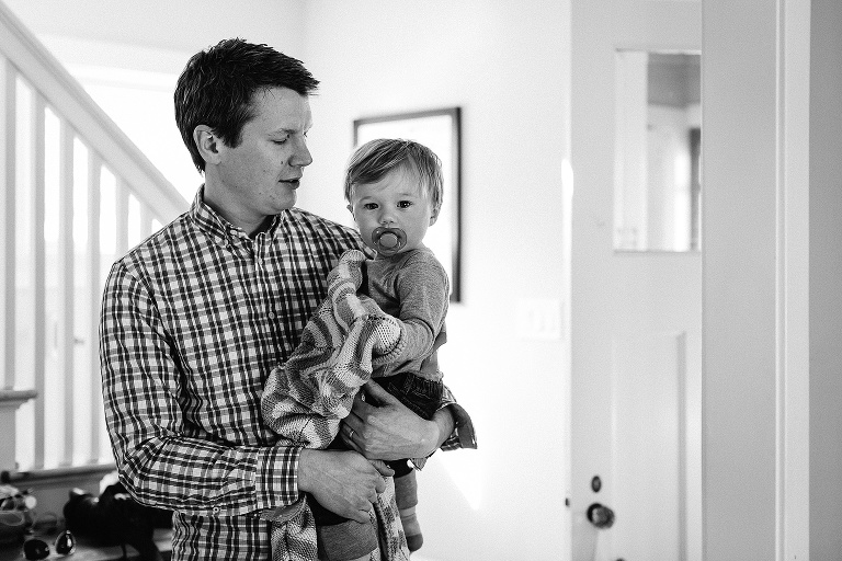Black and white. Father holds toddler with binkie and blanket.