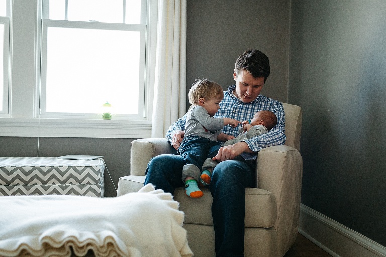 Father sits in chair with toddler son and newborn baby.