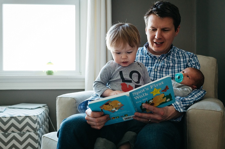 Father sitting in a chair reads story to toddler and newborn baby.