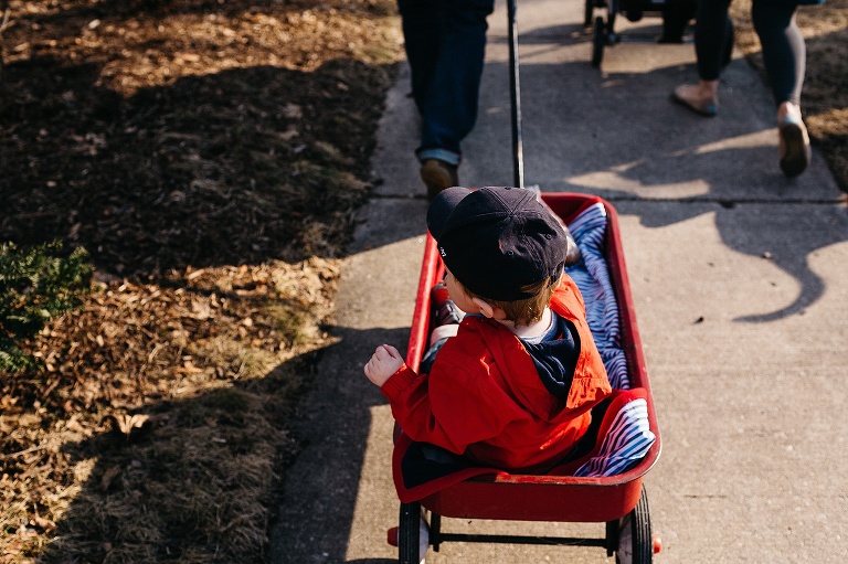 Mother pushes a stroller with newborn baby and father pulls a wagon with toddler down the sidewalk.