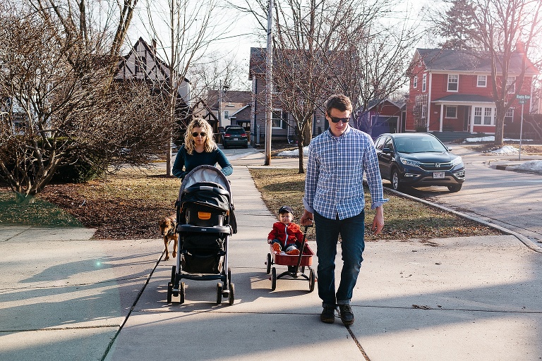 Family walks down the sidewalk with stroller, wagon, and dog.
