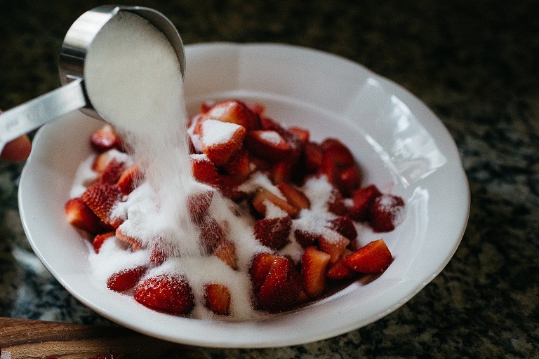 White sugar being poured onto a bowl of fresh sliced strawberries