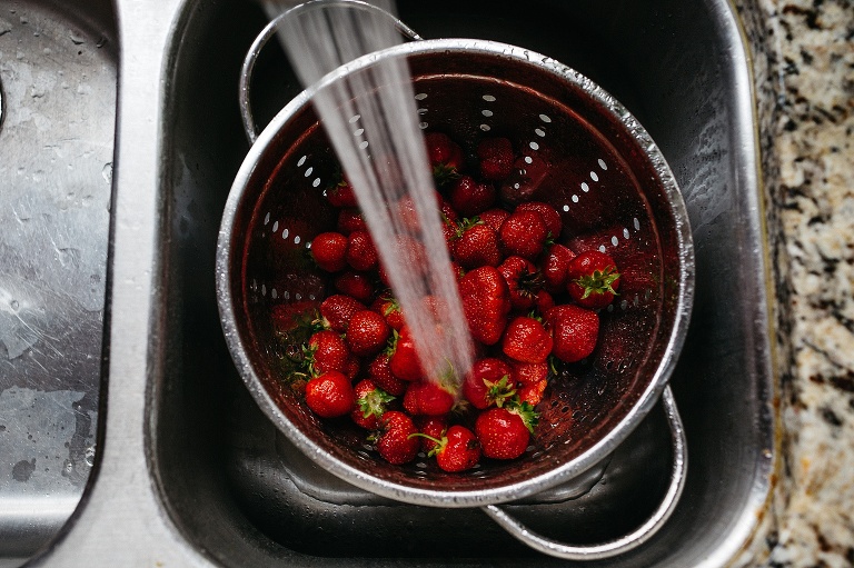 Colander full of strawberry's sitting in a sink with water running washing them off.