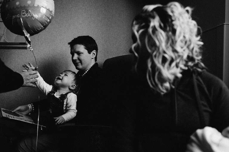 Black and white. Family in hospital room welcoming newborn baby boy. Father with son on his lap holding baby balloons.