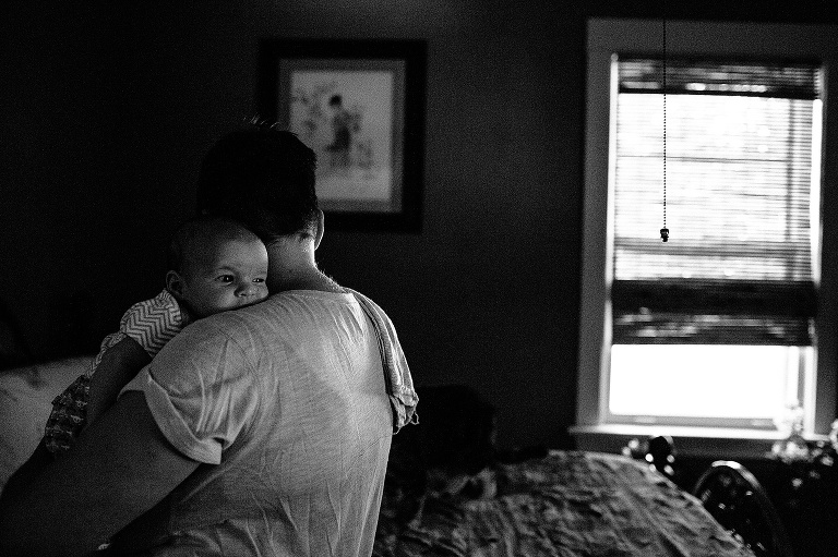 Black and white. Woman in her bedroom holds baby boy as he looks over her shoulder.
