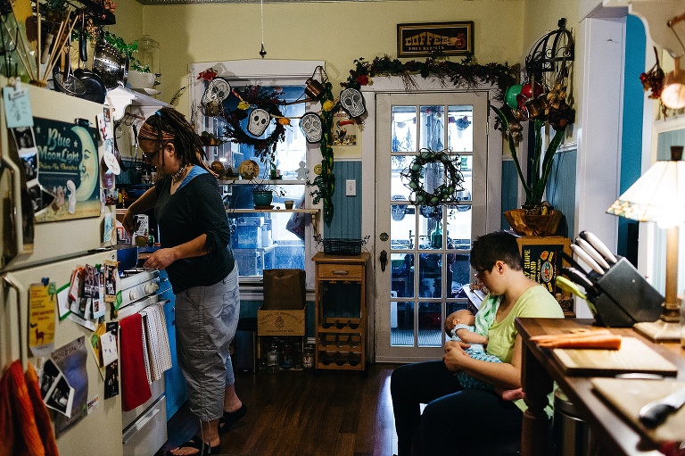 Two woman in a blue and yellow kitchen, one cooking on the stove, the other sitting and holding sleeping newborn baby.