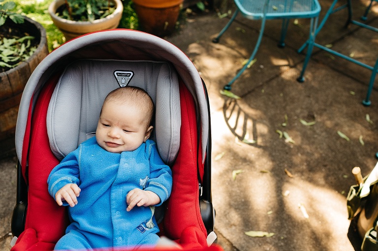 Baby boy wearing blue fuzzy onesie lays in red car seat stroller on backyard patio.