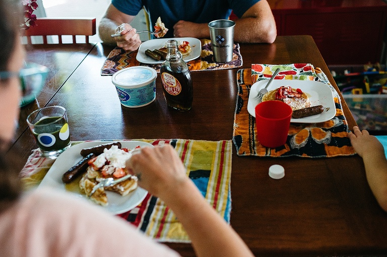 Family sits at kitchen table eating a breakfast of strawberry pancakes and sausages.