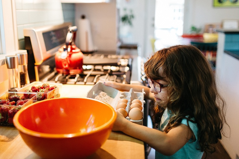 Young girl helps make pancakes in the kitchen. She opens a box of eggs getting ready to crack it in the orange mixing bowl.