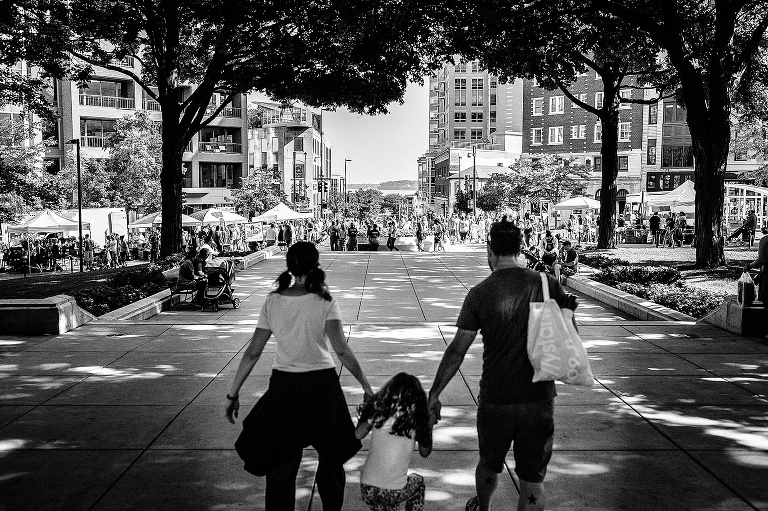 Black and white. Mother and father hold hands with their daughter as they walk down a path to a farmers market.