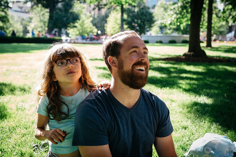 Father and daughter sit in the grass looking up at the sky.