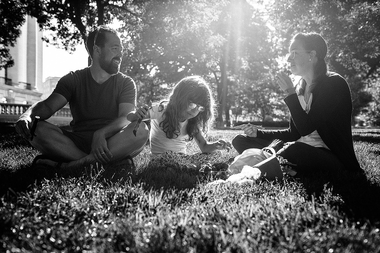 Black and white. Mother, father and daughter sit in the grass eating beef jerky at a farmers market.