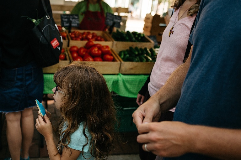 Location: Farmers market. Young girl eating a blue cookie, mother and father walking behind her