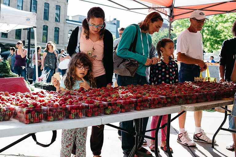 Mother and daughter at a farmers market stand in front of a table full of strawberry's picking one out.
