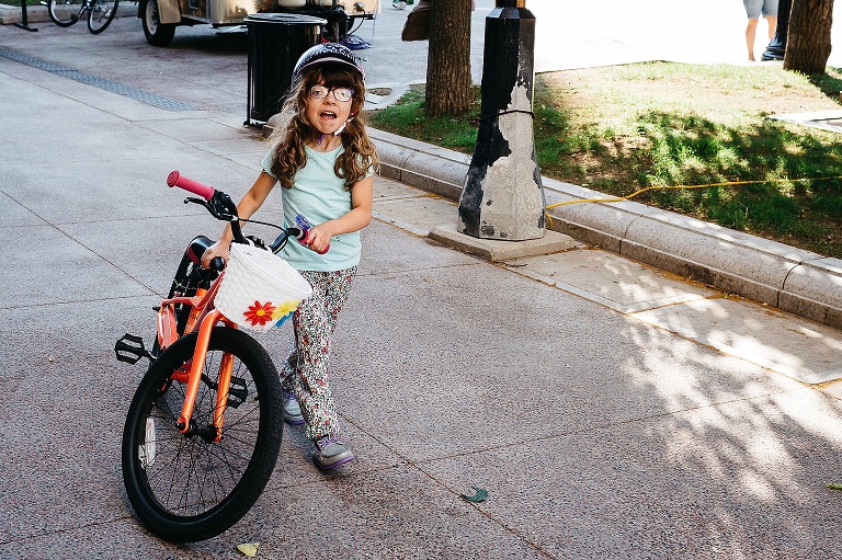 Young girl with curly brown hair and wearing helmet walks her orange bike down a path.