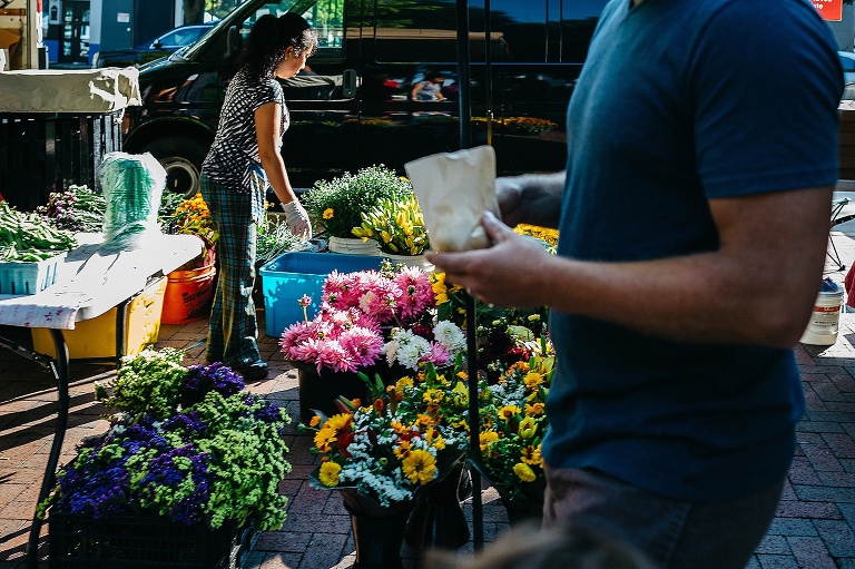 Man walking at a famers market. woman working at a flower stand is behind him.