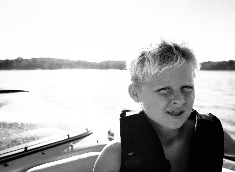 Black and white. Young boy wearing a life jacket sitting on a boat in the middle of the lake.