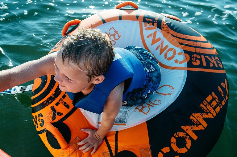 Kid on a inter tube on a lake climbs back into boat.
