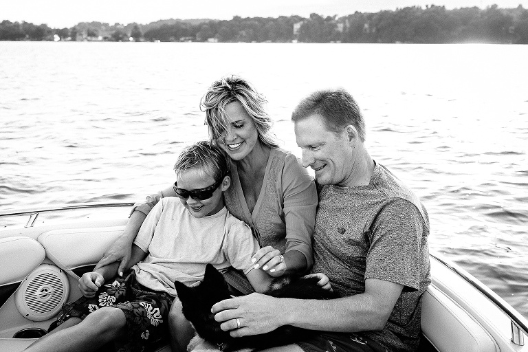 Black and white. Mother, father, son, and puppy sit on motorboat.