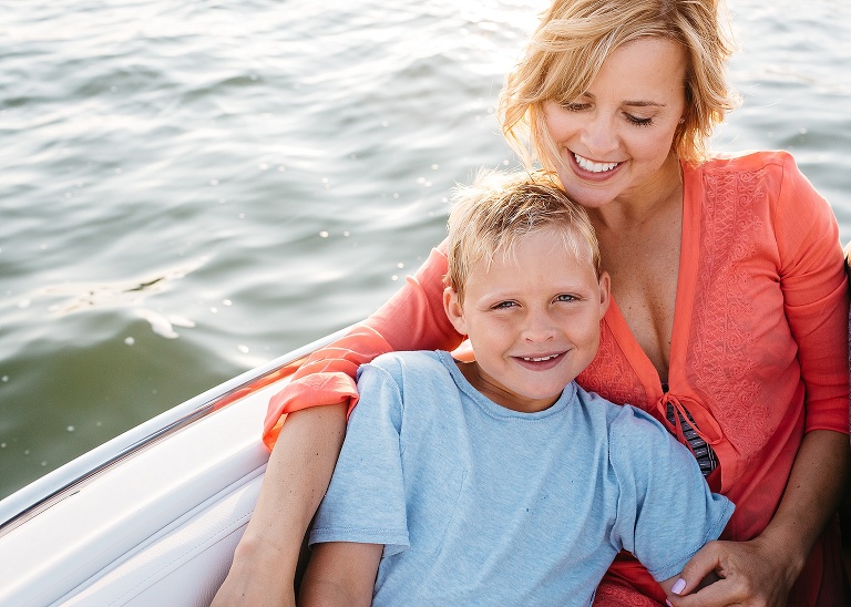 Mother and son sit in a motorboat on the lake together. Moms arm wrapped around her son.