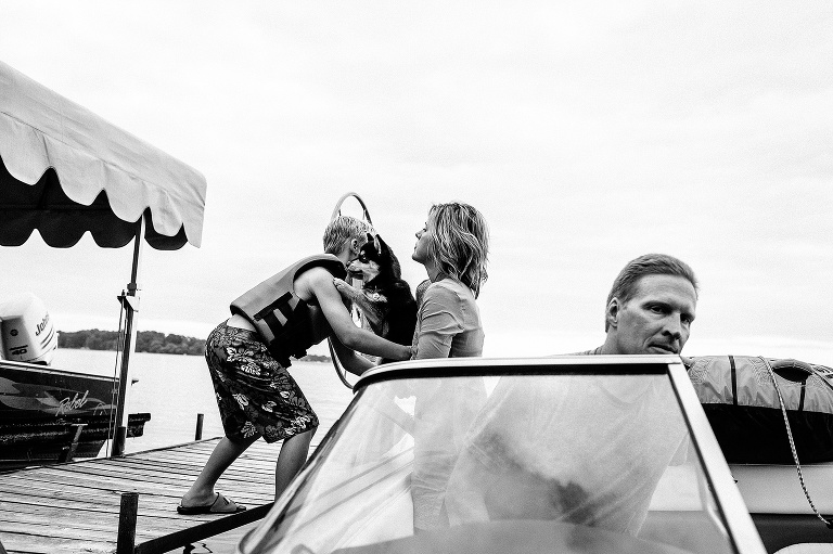 Black and white. Mother on boat hands puppy to her son standing on a dock.