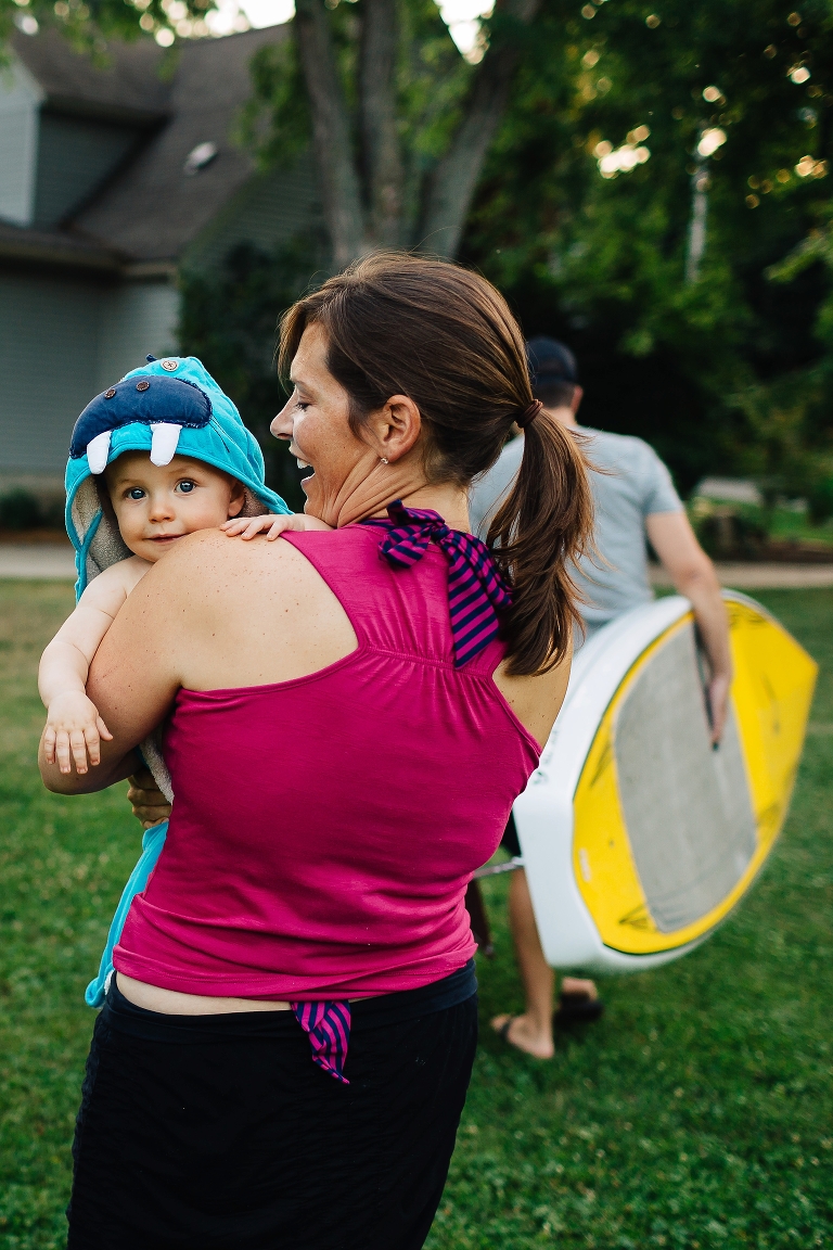 Dad carries stand up paddle board while mom carries baby son wearing blue monster town over his head.