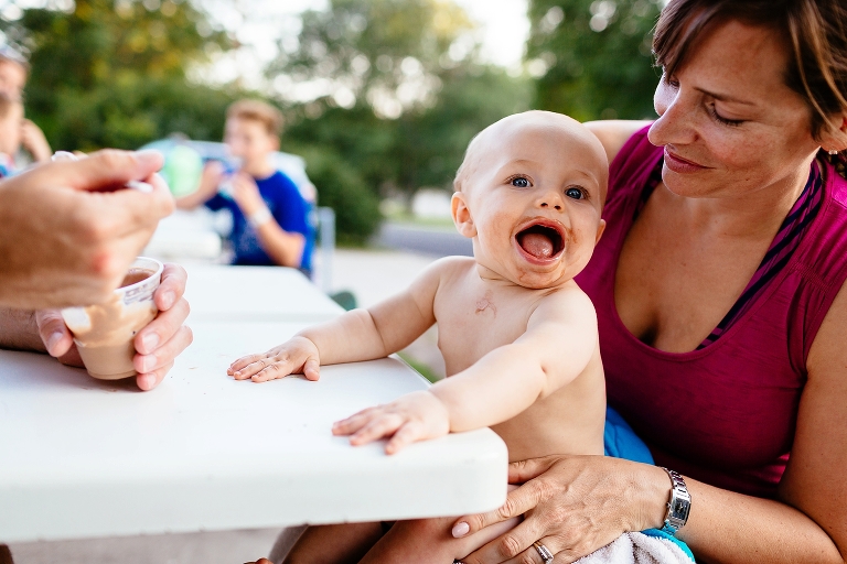 Baby sitting on mothers lap smiles with chocolate ice cream around his mouth.