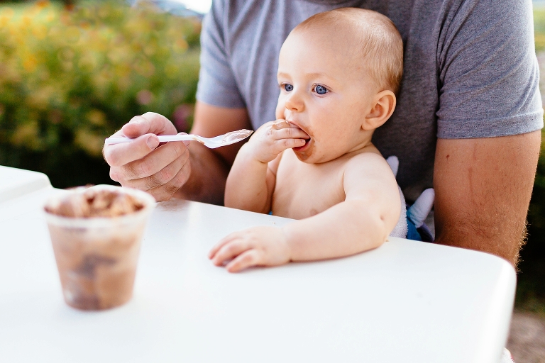 Baby sits on fathers lap as the two share a bowl of chocolate ice cream.