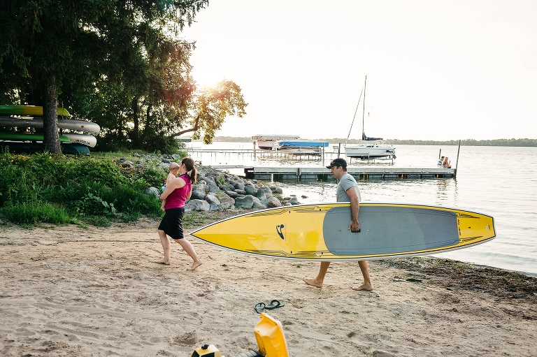 Mother holds onto baby boy and father carries the big yellow stand up paddle board as they walk through the sand.