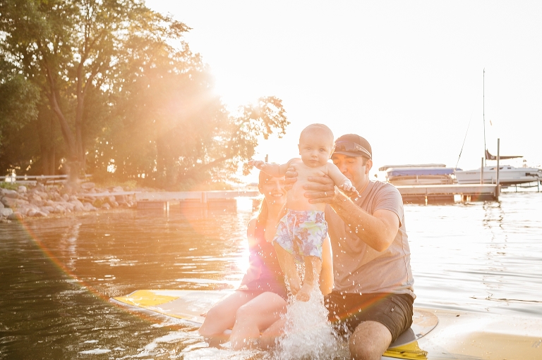 Mother and father sitting on stand up paddle board pick up son splashing water below him. Sun setting behind them shining into the camera.