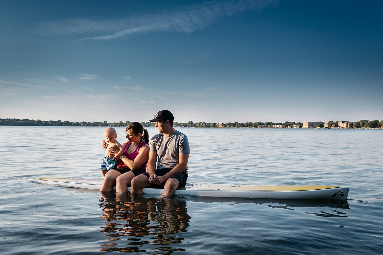 Mother and father sitting sideways on a SUP feet dangling into the water holding onto their baby son. Beautiful blue sky behind them.