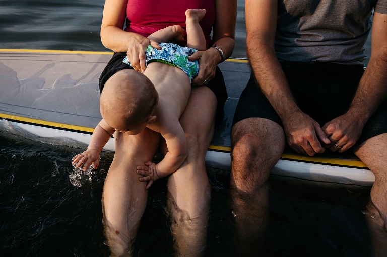 Mother and father sitting sideways on a SUP feet dangling into the water holding onto their baby son.