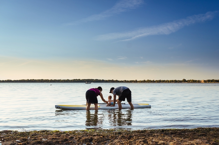 Mother and father hold onto baby sitting on a stand up paddle board in the lake