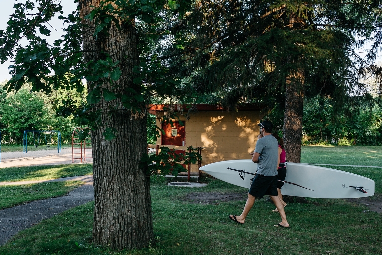 A couple walks across park grass carrying a stand up paddle board.