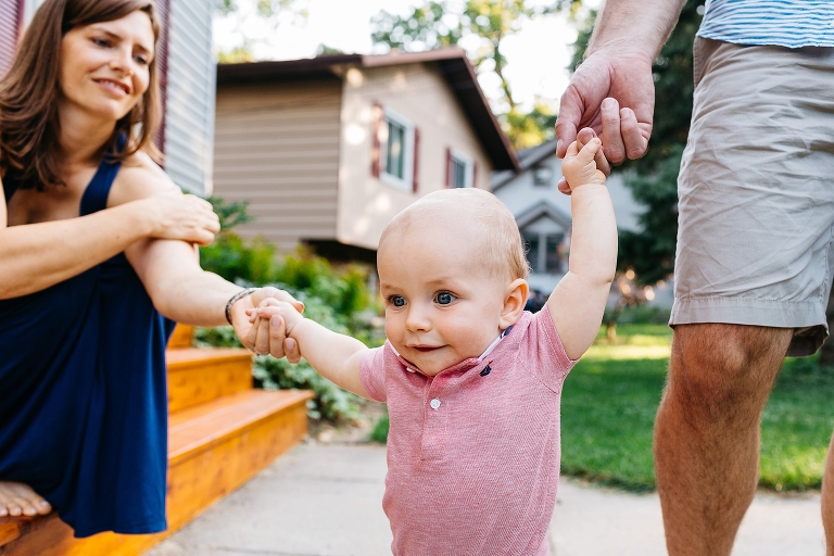 Mother and father hold baby's hands helping him walk.