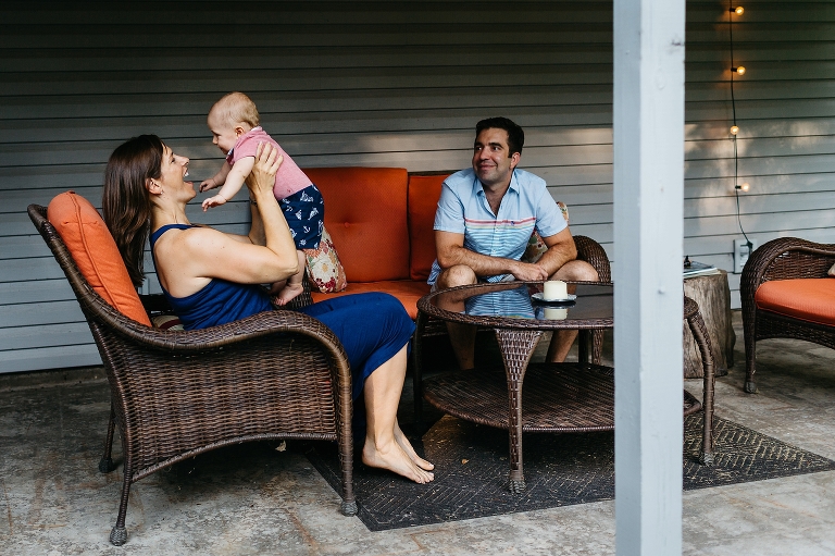 Mother sitting in patio hair holds up her baby boy, father watching from the couch.