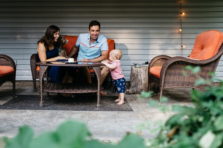 Mother and father sit on patio couch watching son stand up holding onto patio table.