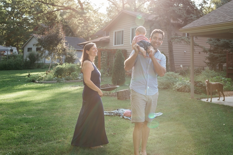 Father standing in his backyard carrying baby boy on his shoulder, Mom looking from behind and dog watching from the patio.
